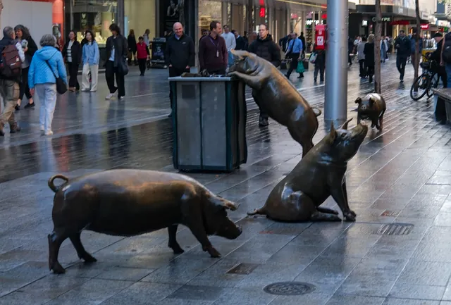 Three of the four bronze pigs among shoppers in Rundle Mall, one with its front feet up on a rubbish bin