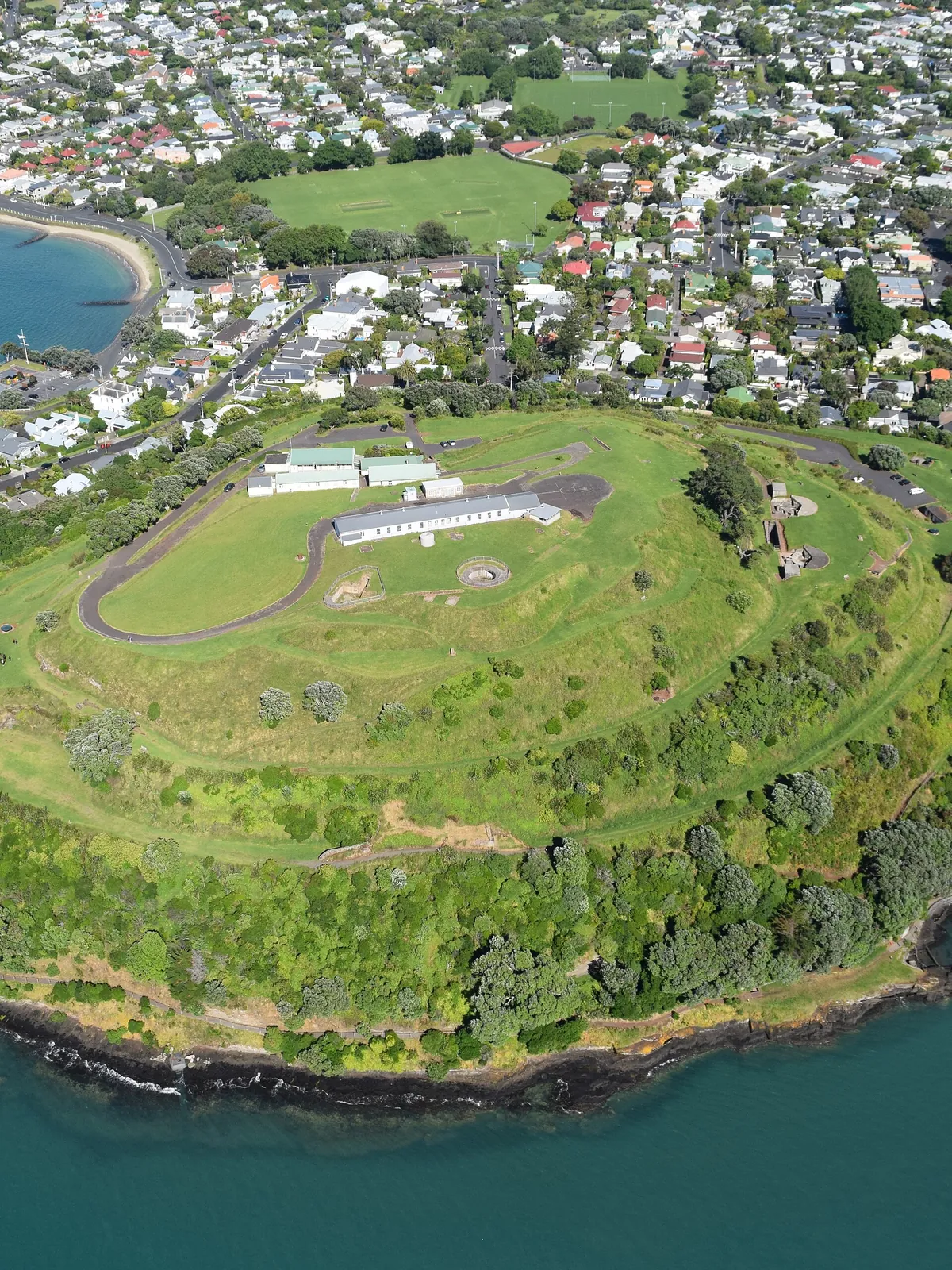 Maungauika, North Head, from the air: a green volcanic headland with its summit road, Devonport's houses and beaches behind