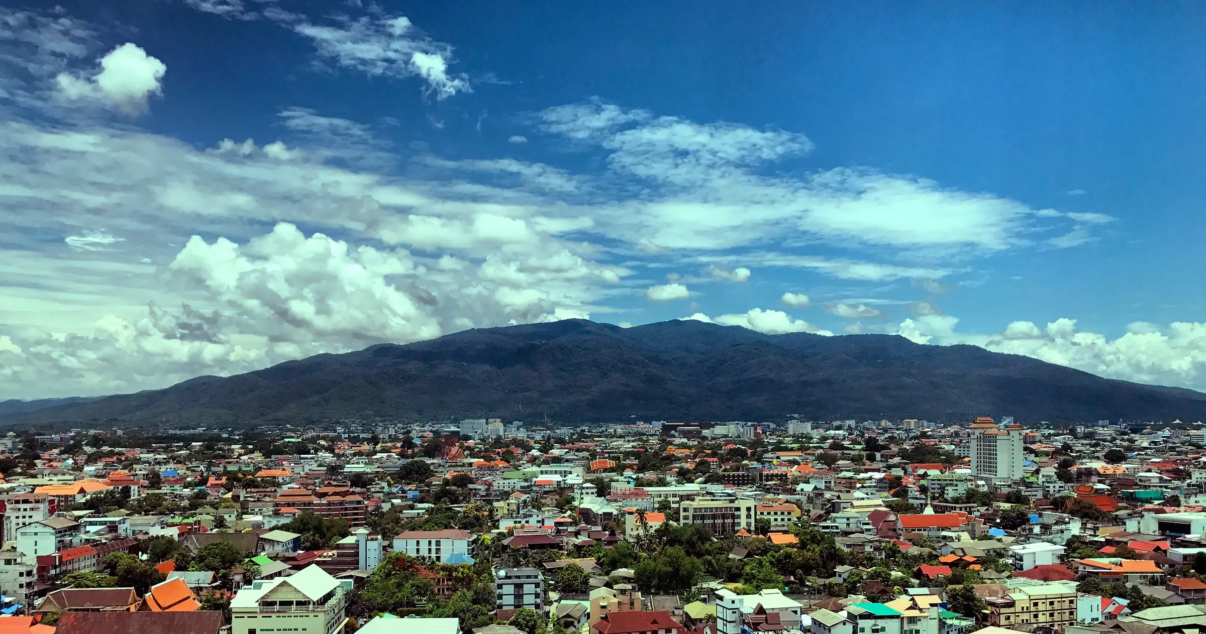 Chiang Mai from above on a bright day: a sea of red, orange and blue roofs and green trees running to the long, dark ridge of Doi Suthep under a blue sky full of white clouds