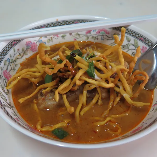 A bowl of khao soi: curry noodle soup topped with a tangle of crisp fried noodles, a china spoon and chopsticks on the rim