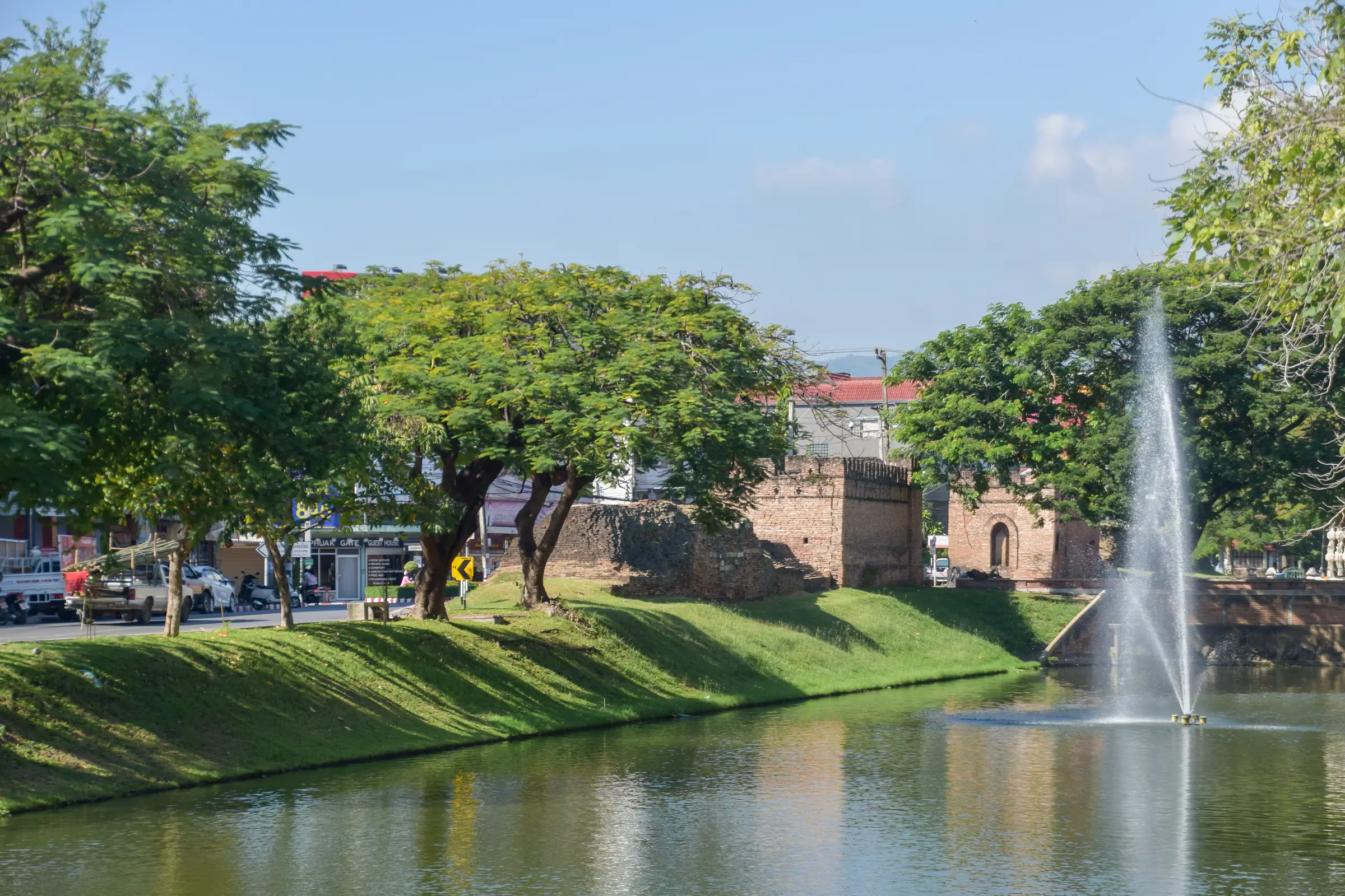 The old city moat on a sunny morning: a fountain playing in the green water, grassy banks and big shade trees, the brick walls of a gate beyond