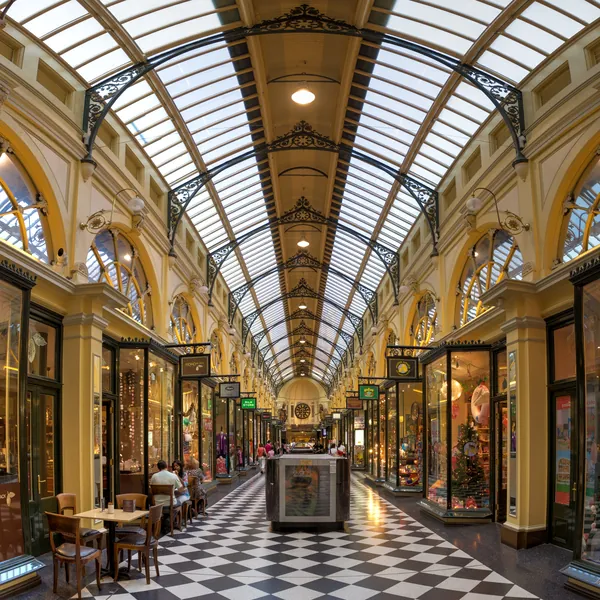 Inside the Royal Arcade: a long Victorian shopping arcade under an arched glass roof, its shopfronts lit gold and its floor laid in black and white squares
